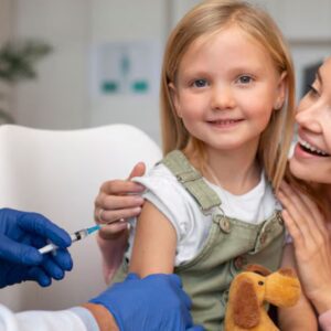 Bebê sorrindo com um adesivo de curativo no braço após vacinação, com uma enfermeira sorrindo ao fundo, simbolizando a proteção da vacinação infantil.