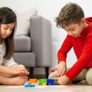Duas crianças pequenas concentradas brincando juntas com blocos coloridos no chão de uma sala de estar, com um sofá ao fundo, simbolizando o impacto positivo do desenvolvimento infantil e o uso saudável de telas ao priorizar brincadeiras interativas.
