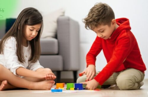 Duas crianças pequenas concentradas brincando juntas com blocos coloridos no chão de uma sala de estar, com um sofá ao fundo, simbolizando o impacto positivo do desenvolvimento infantil e o uso saudável de telas ao priorizar brincadeiras interativas.